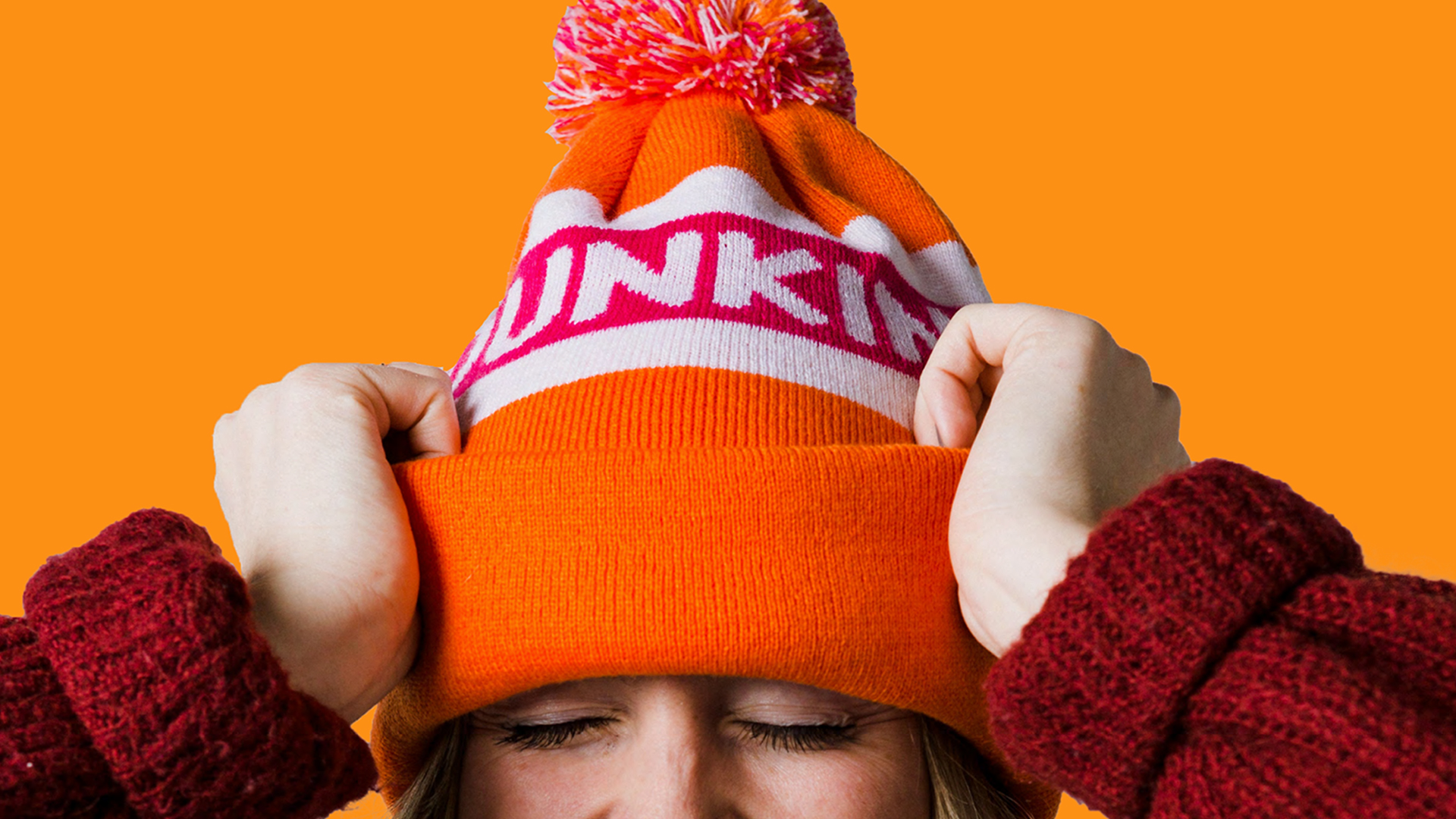 A close up of a person wearing an orange and pink Dunkin' beanie with a pom pom on top