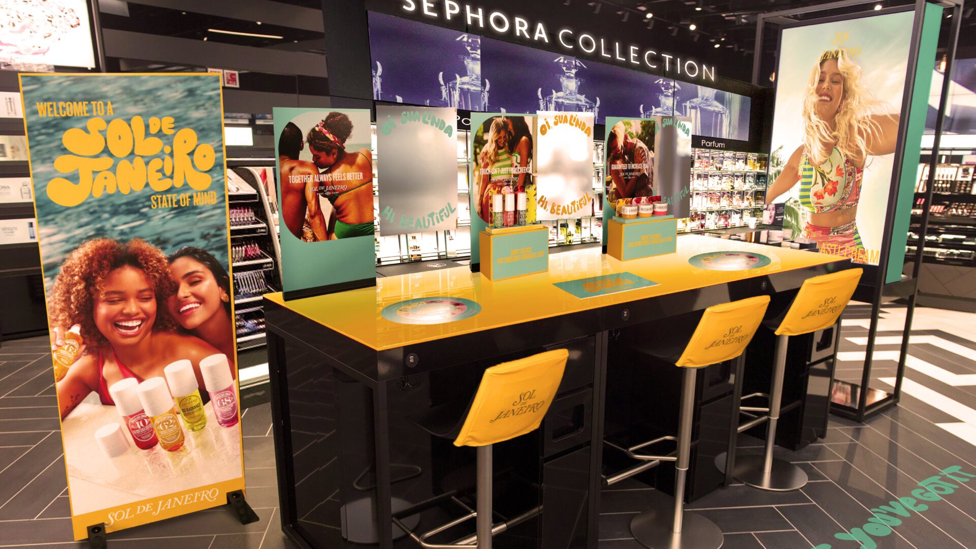 A Sol De Janeiro makeup counter in a sephora store. Freestanding banner on the left, mirror and counter graphics in the center, chairs with branded chair covers, and and branded graphics on the floor.